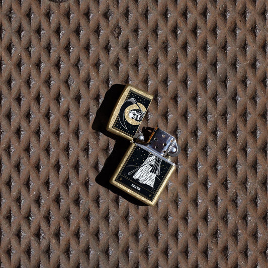 Lifestyle image of Zippo Death Card Design Street Brass Windproof Lighter with its lid open and unlit laying on a rusted diamond tread surface.