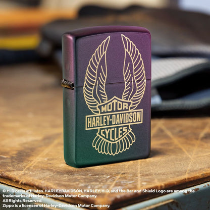 Lifestyle image of Zippo Harley-Davidson® Winged Logo Design Iridescent Windproof Lighter standing at a 3/4 angle next to work gloves on a workbench showing the front and hinge side of the lighter. 

