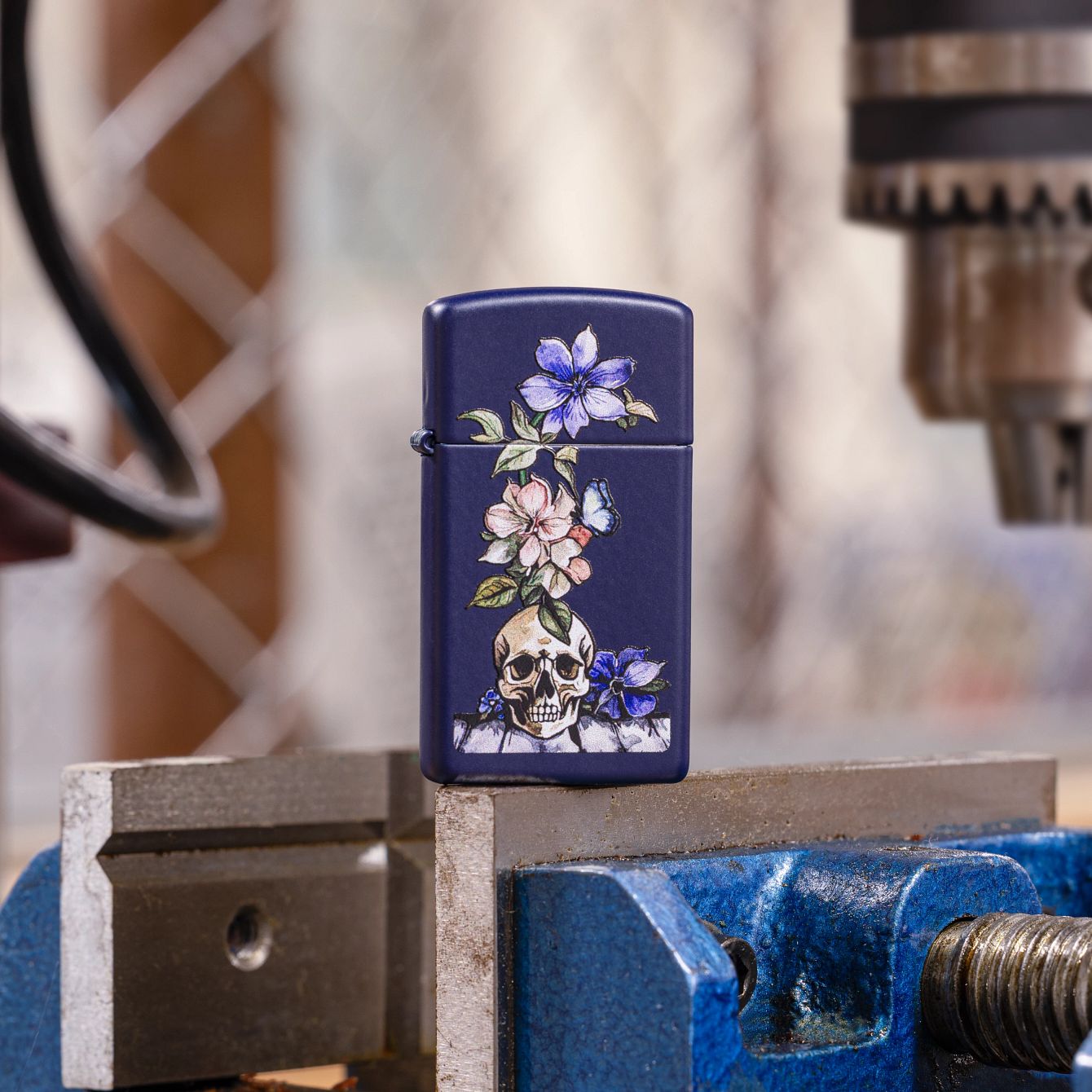 Lifestyle image of Zippo Bone Bouquet Design Slim Navy Matte Windproof Lighter standing on top of a blue and silver workshop vise with a blurred workshop as the background. 

